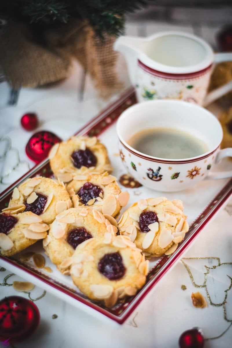 A festive holiday tray filled with raspberry almond thumbprint cookies topped with sliced almonds and a center of red raspberry jam. The tray sits next to a Christmas-themed cup of coffee and cream pitcher, surrounded by red ornaments and a decorated holiday tablecloth, creating a cozy Christmas morning scene.