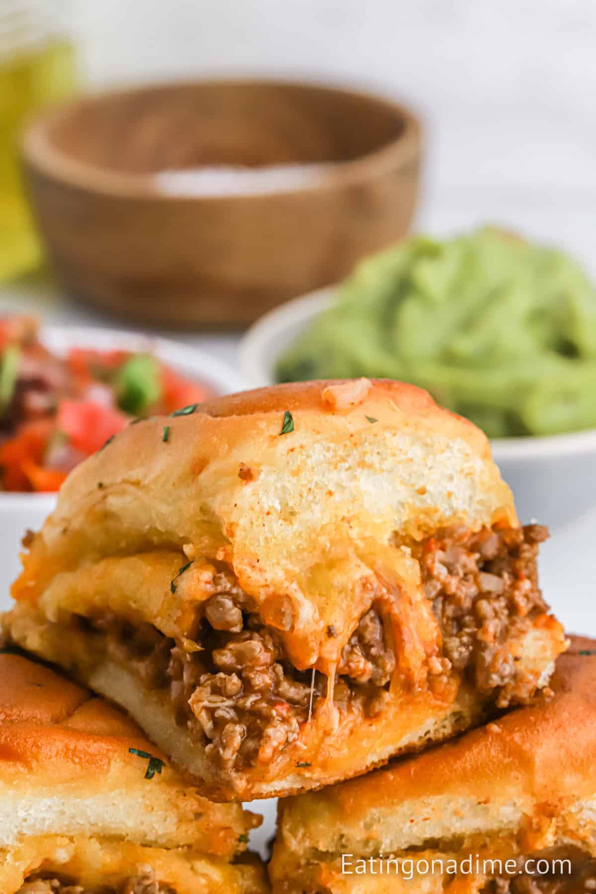 Close-up of a cheesy taco slider made with seasoned ground beef and melted cheese, baked in a soft golden Hawaiian roll. Served with bowls of guacamole and salsa in the background.