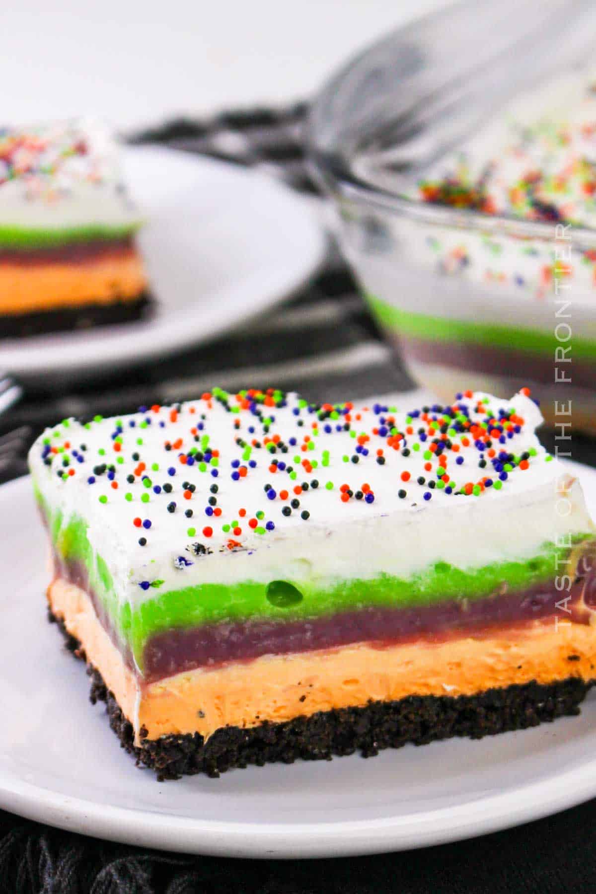 Layered Halloween lush dessert with Oreo crust, orange pudding, green and purple layers, whipped topping, and Halloween sprinkles, served on a plate for a festive and colorful party treat.
