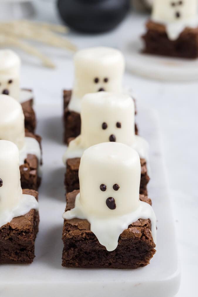 Fudgy brownie squares topped with marshmallows decorated as little white chocolate ghosts with candy eyes and mouths, making a fun and spooky Halloween treat.