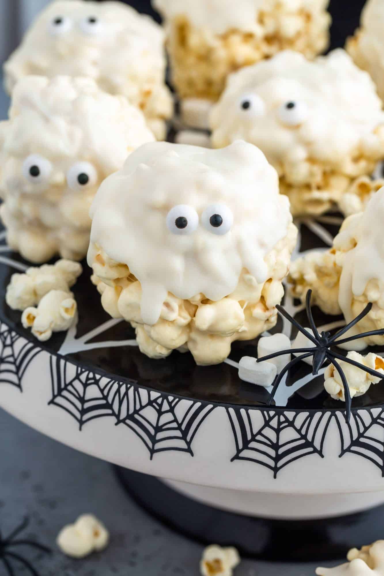 Popcorn balls coated in white chocolate, decorated with candy eyes to look like little ghosts, displayed on a spiderweb cake stand for a fun and spooky Halloween treat.