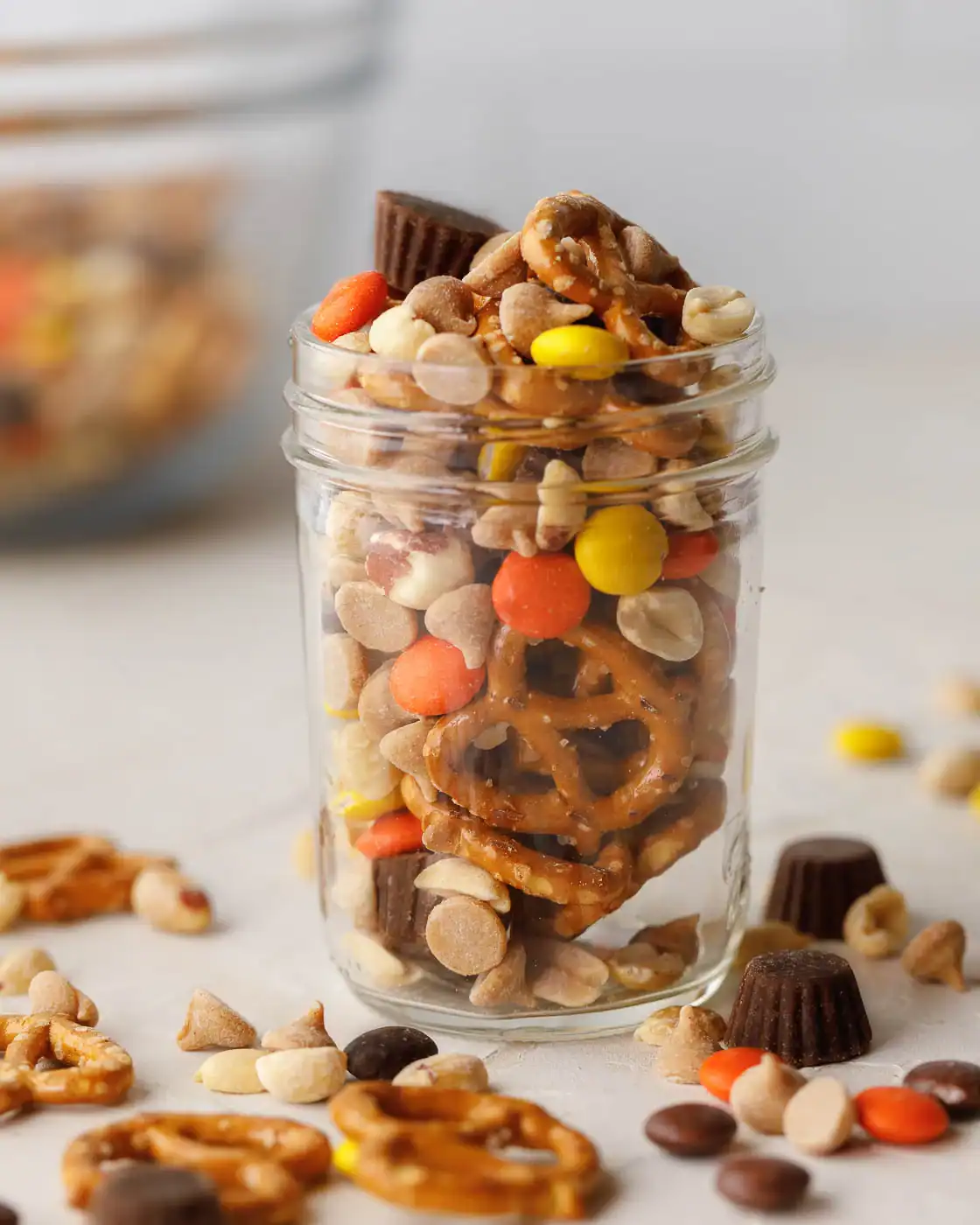 Glass jar filled with Halloween trail mix made of pretzels, candy-coated chocolates, mini peanut butter cups, peanuts, and butterscotch chips, with extra pieces scattered around for a festive party snack.