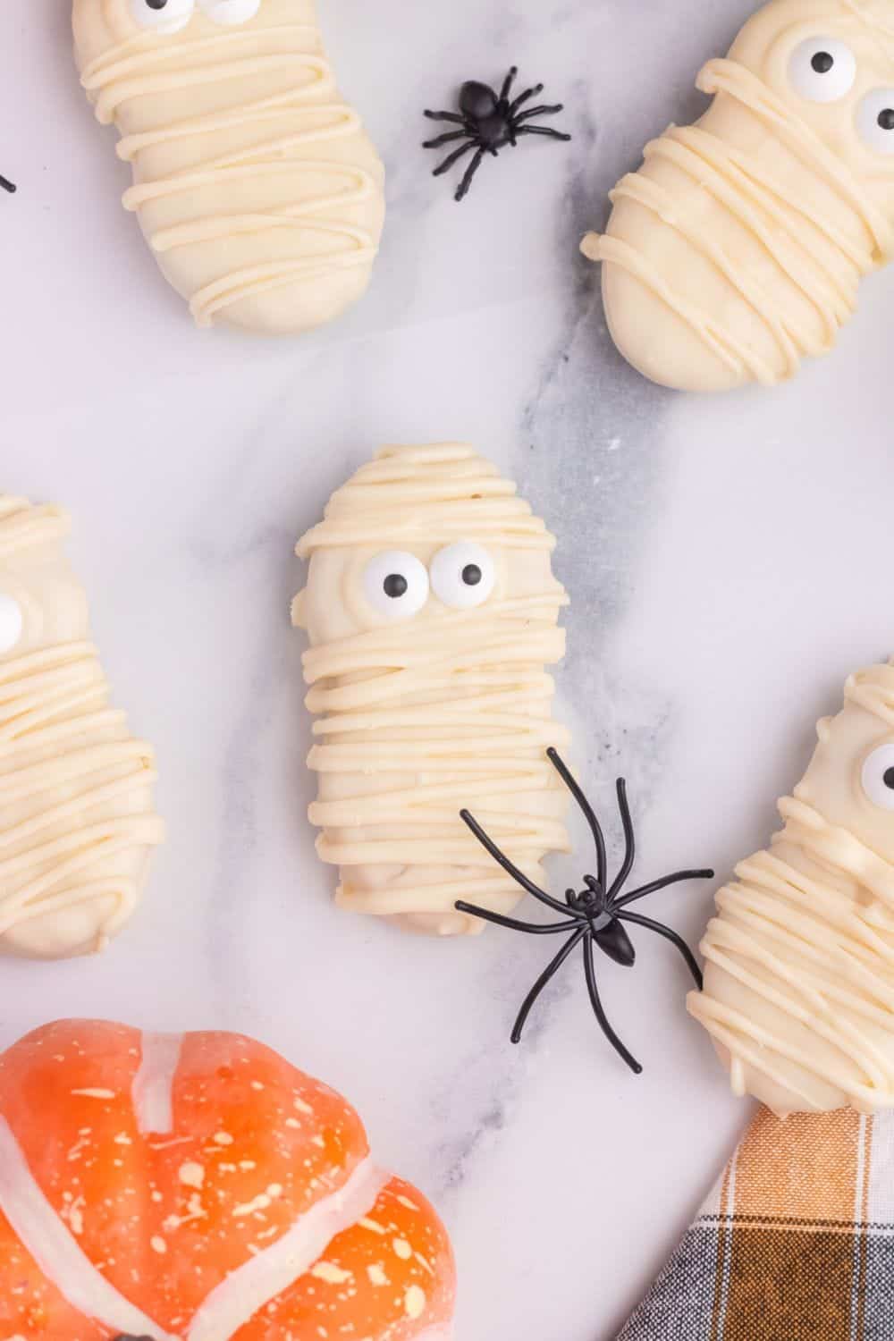 Nutter Butter cookies coated in white chocolate, drizzled with extra chocolate to look like mummy bandages, and decorated with candy eyes, arranged with plastic spiders for a fun Halloween treat.