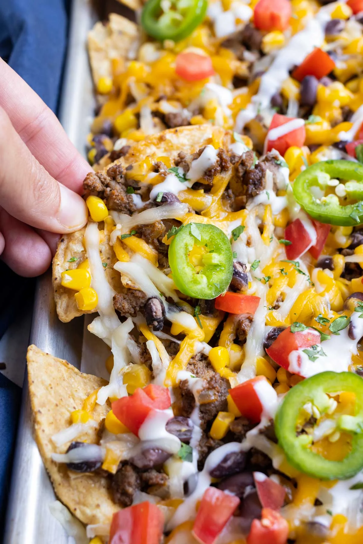 Close-up of loaded sheet pan nachos topped with ground beef, melted cheese, black beans, corn, diced tomatoes, jalapeño slices, and a drizzle of sour cream. A hand is lifting one cheesy chip from the pan.