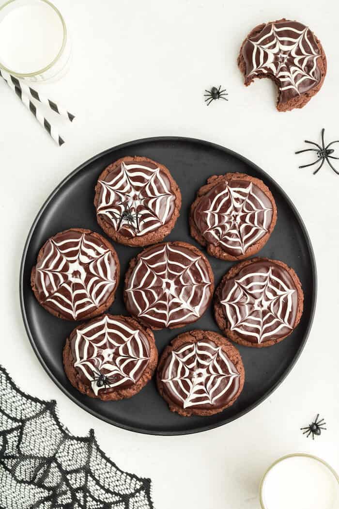 Chocolate cake mix cookies decorated with white icing spiderwebs, arranged on a black plate with toy spiders, making a festive and spooky Halloween dessert.
