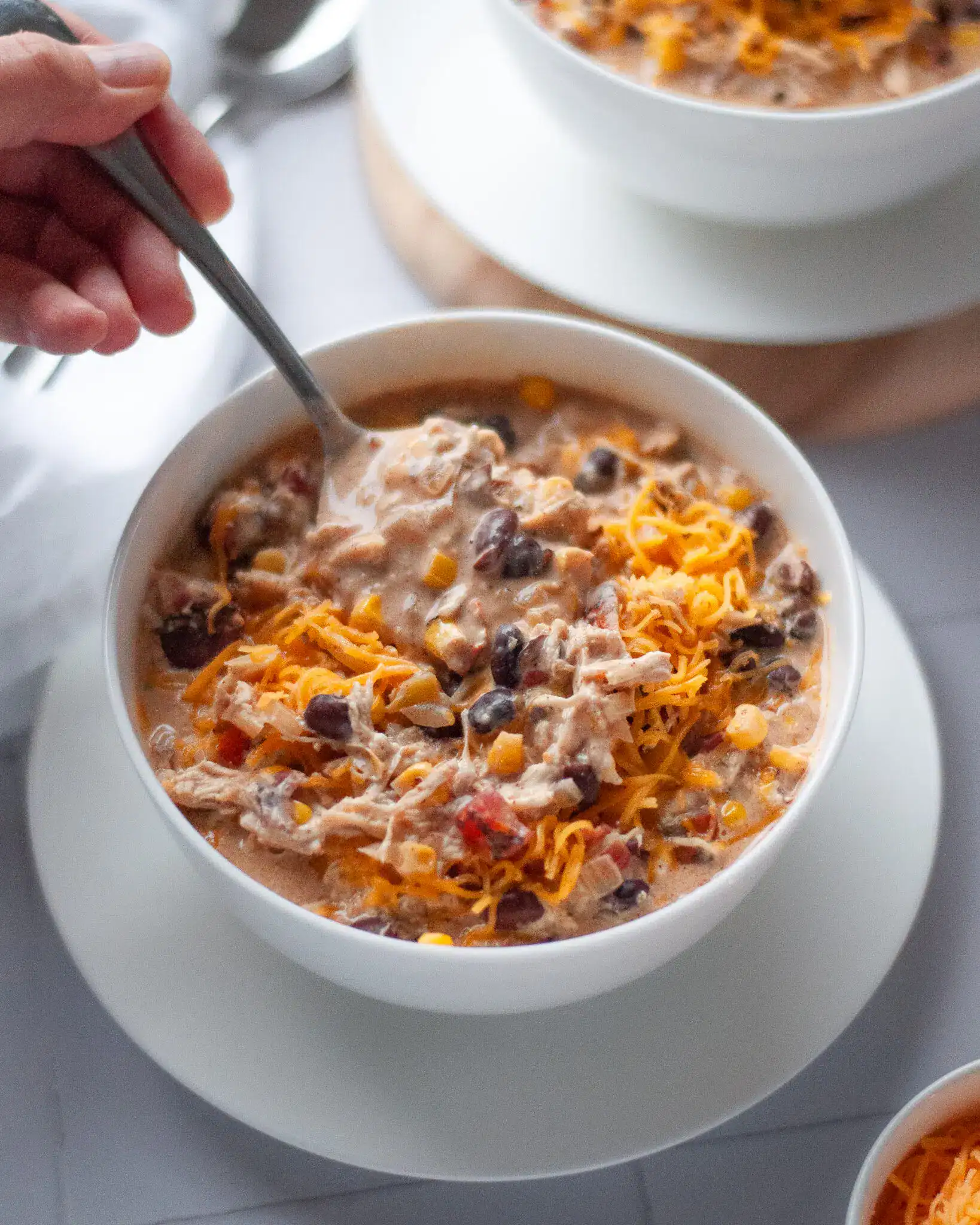 A bowl of creamy white chicken chili topped with shredded cheddar cheese, filled with black beans, corn, shredded chicken, and diced peppers, with a spoon scooping from the bowl.
