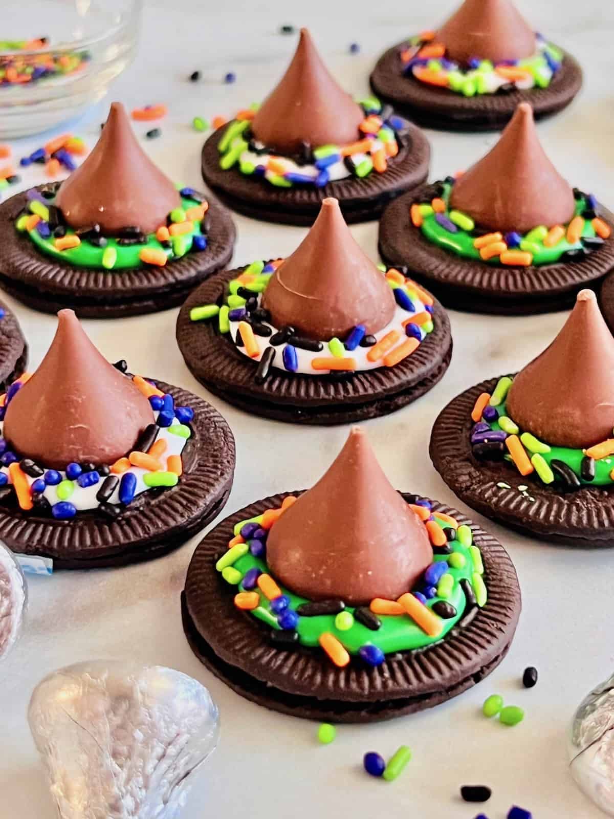 Chocolate Oreo cookies topped with chocolate kisses, colorful Halloween sprinkles, and frosting to look like witch hats, arranged on a counter for a fun and festive Halloween dessert.