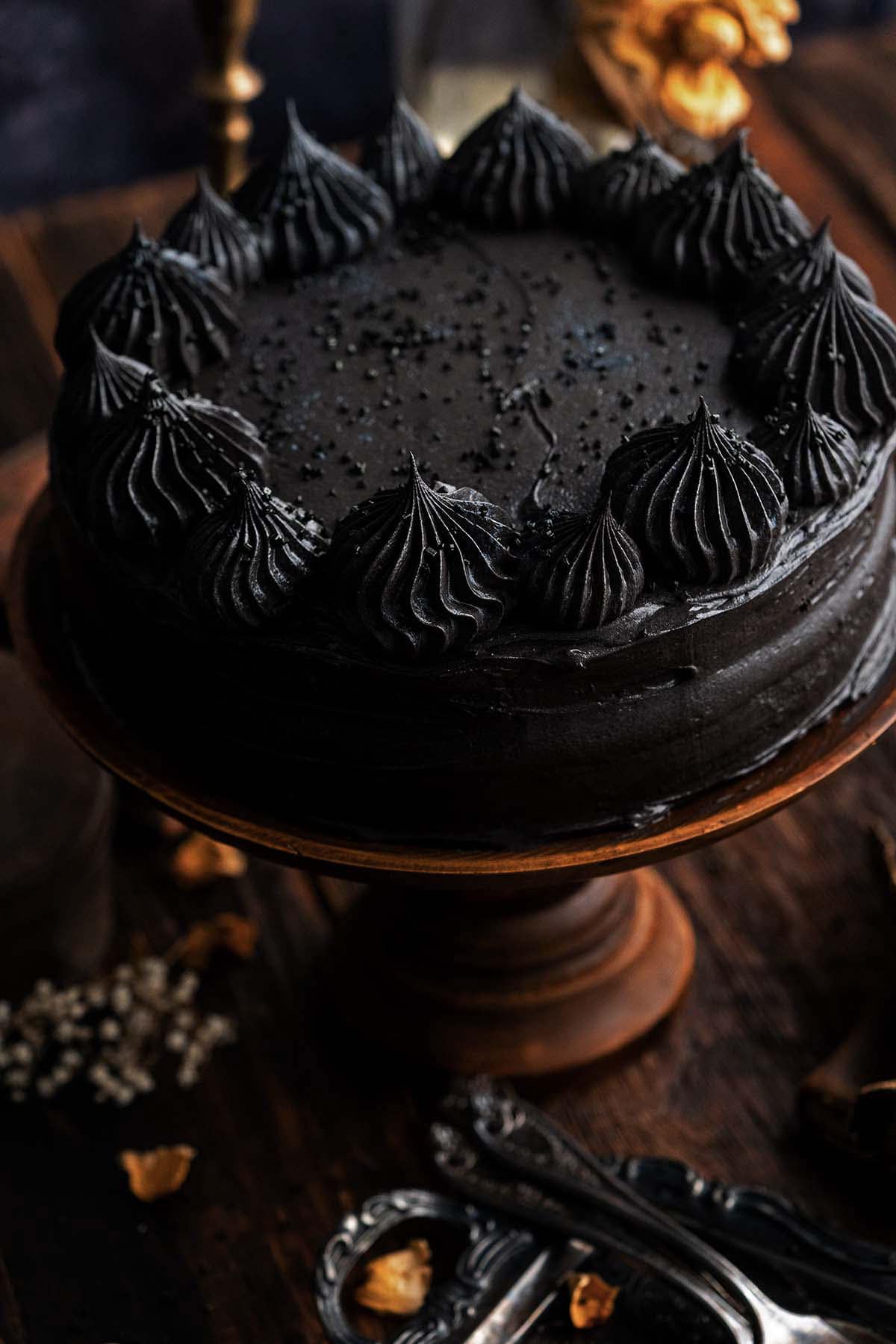 Black cocoa cake with dark black frosting and piped swirls around the edge, topped with black sprinkles, displayed on a wooden cake stand for a dramatic Halloween dessert.