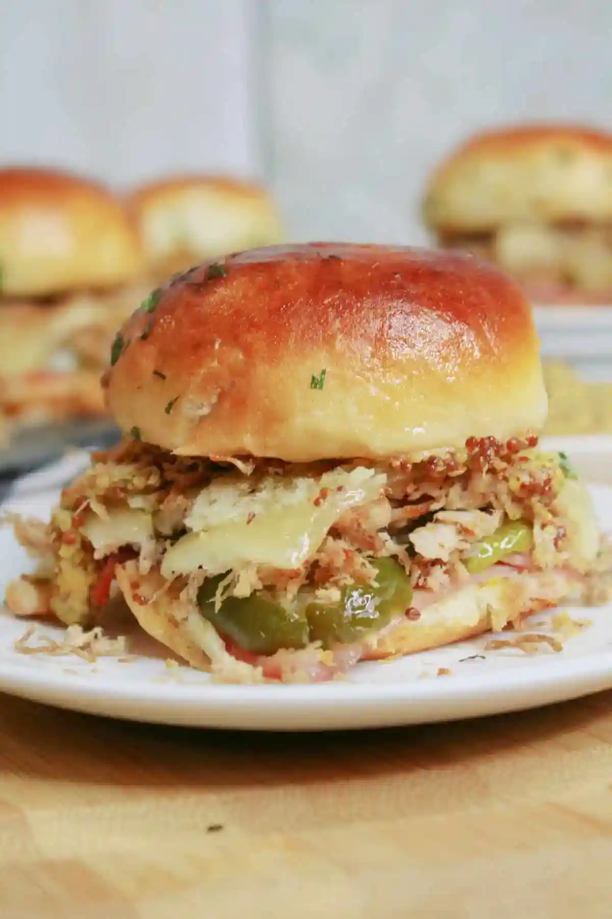 Close-up of a Cuban slider sandwich filled with shredded pork, melted cheese, pickles, and mustard on a golden, buttery roll. Served on a white plate with more sliders in the background.