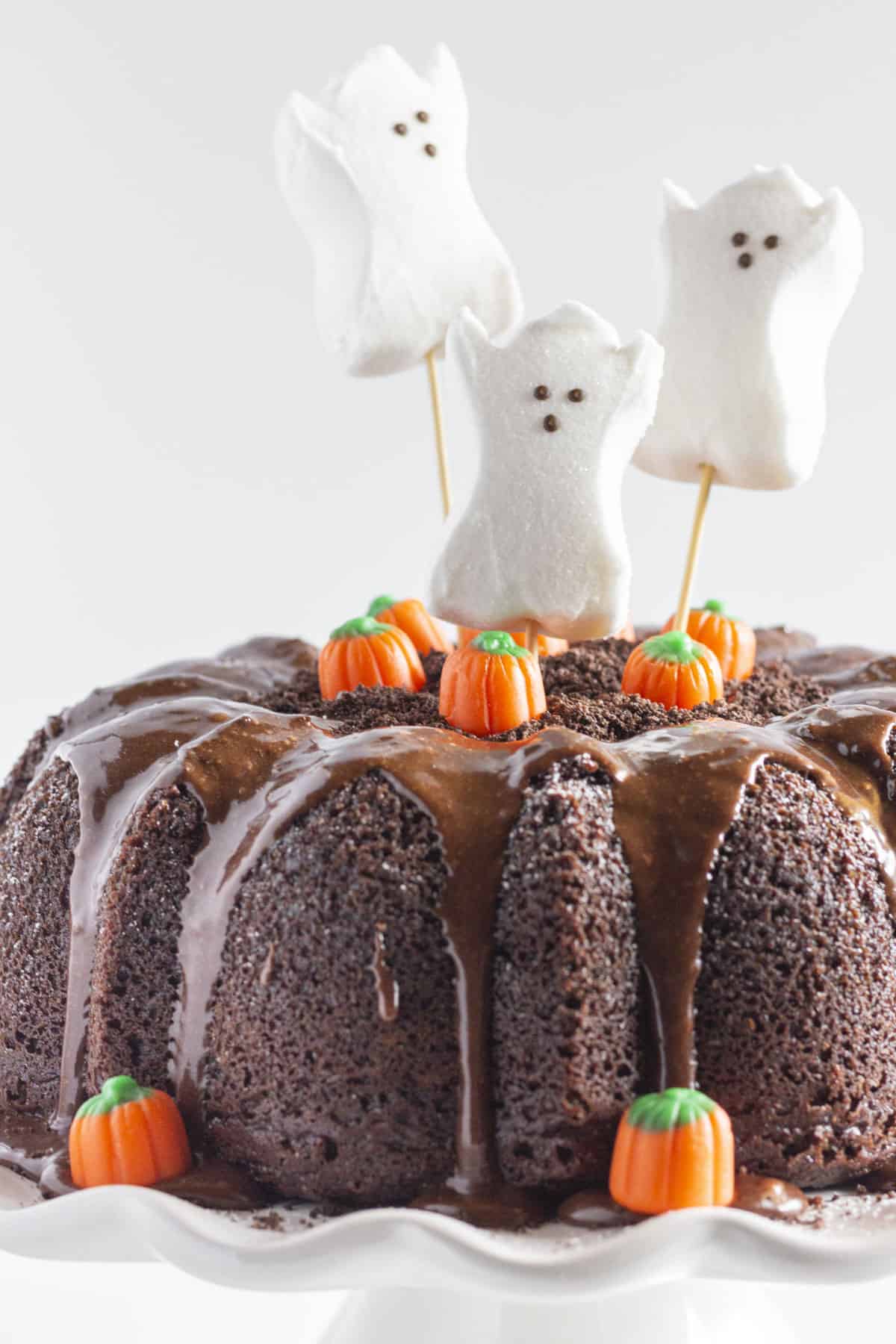 Chocolate bundt cake drizzled with glaze, decorated with marshmallow ghost toppers and candy pumpkins, making a festive and spooky Halloween dessert centerpiece.