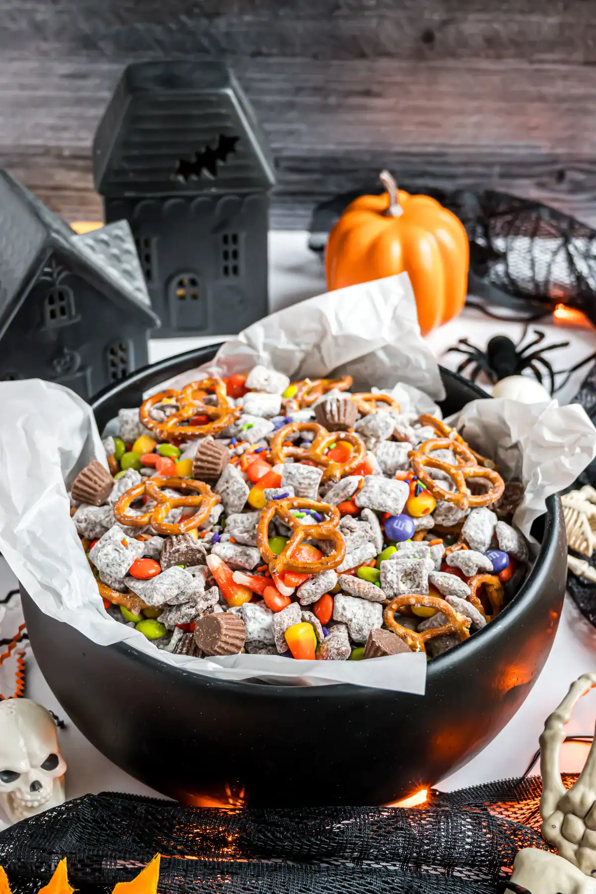 Black cauldron bowl filled with Halloween puppy chow snack mix made of powdered sugar–coated cereal, pretzels, candy corn, chocolate candies, and mini peanut butter cups, surrounded by Halloween decorations.