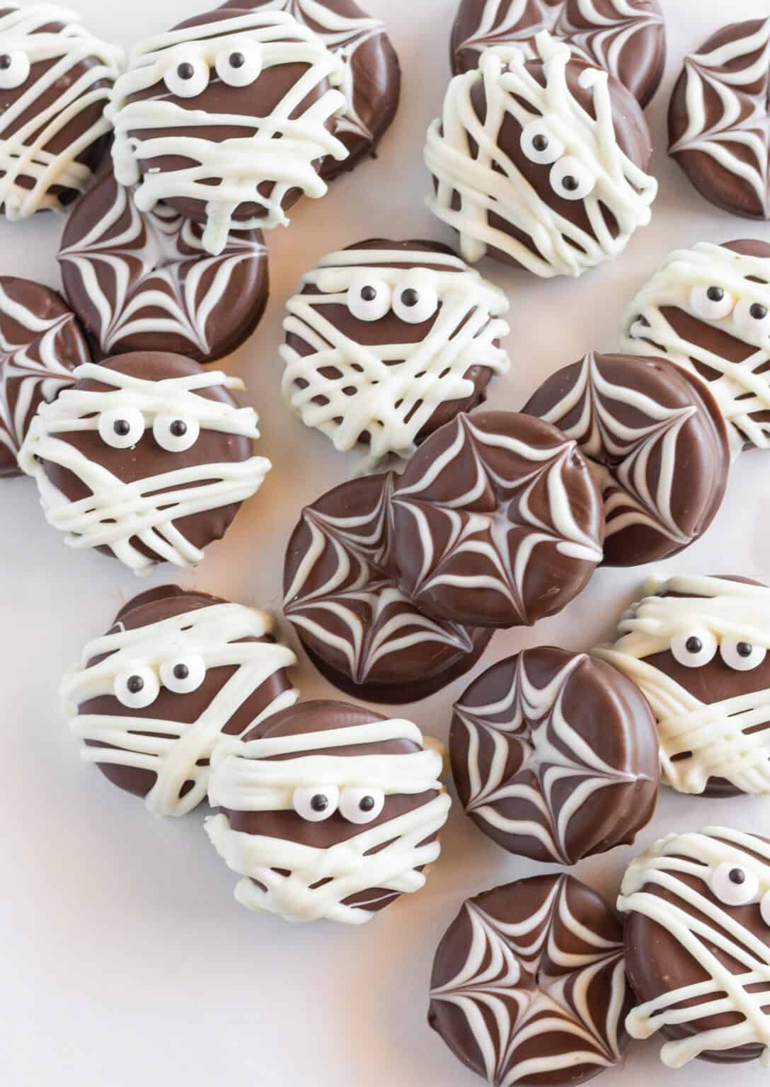 Chocolate-covered Oreo cookies decorated for Halloween with white icing spiderwebs and mummy designs, complete with candy eyes, creating fun and spooky treats perfect for parties and kids.