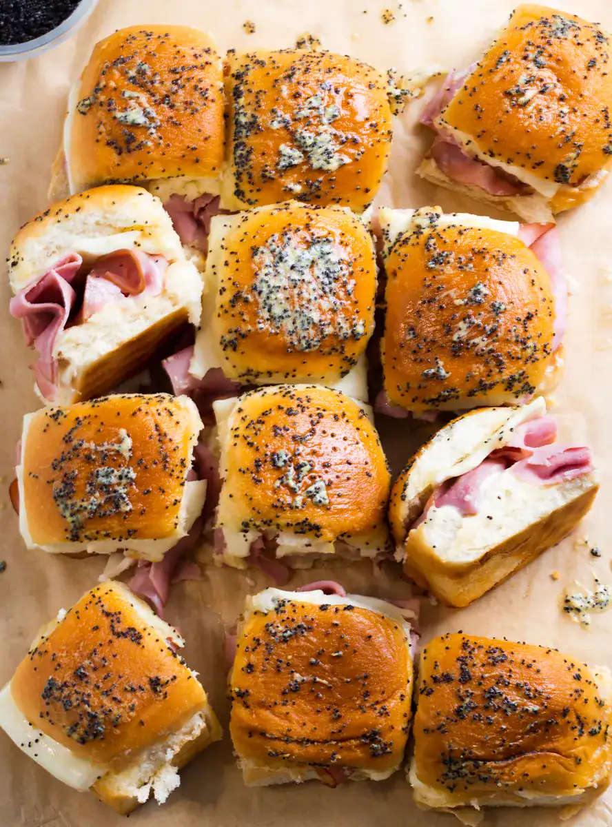 Baked ham and cheese sliders made with soft Hawaiian rolls, topped with poppy seeds and a buttery glaze. The sliders are filled with layers of sliced ham and melted cheese, arranged on parchment paper and ready to serve.