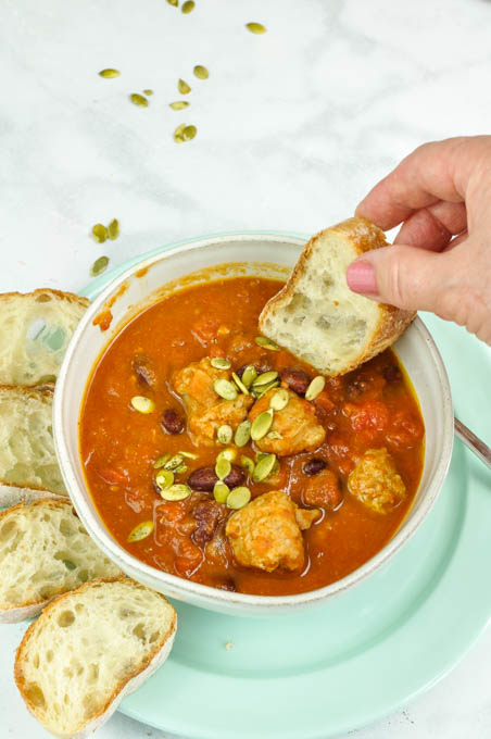 the best Pumpkin Chili with turkey sausage in a white bowl with rounds of bread in a breen plate and a hand dipping in a slice of bread.