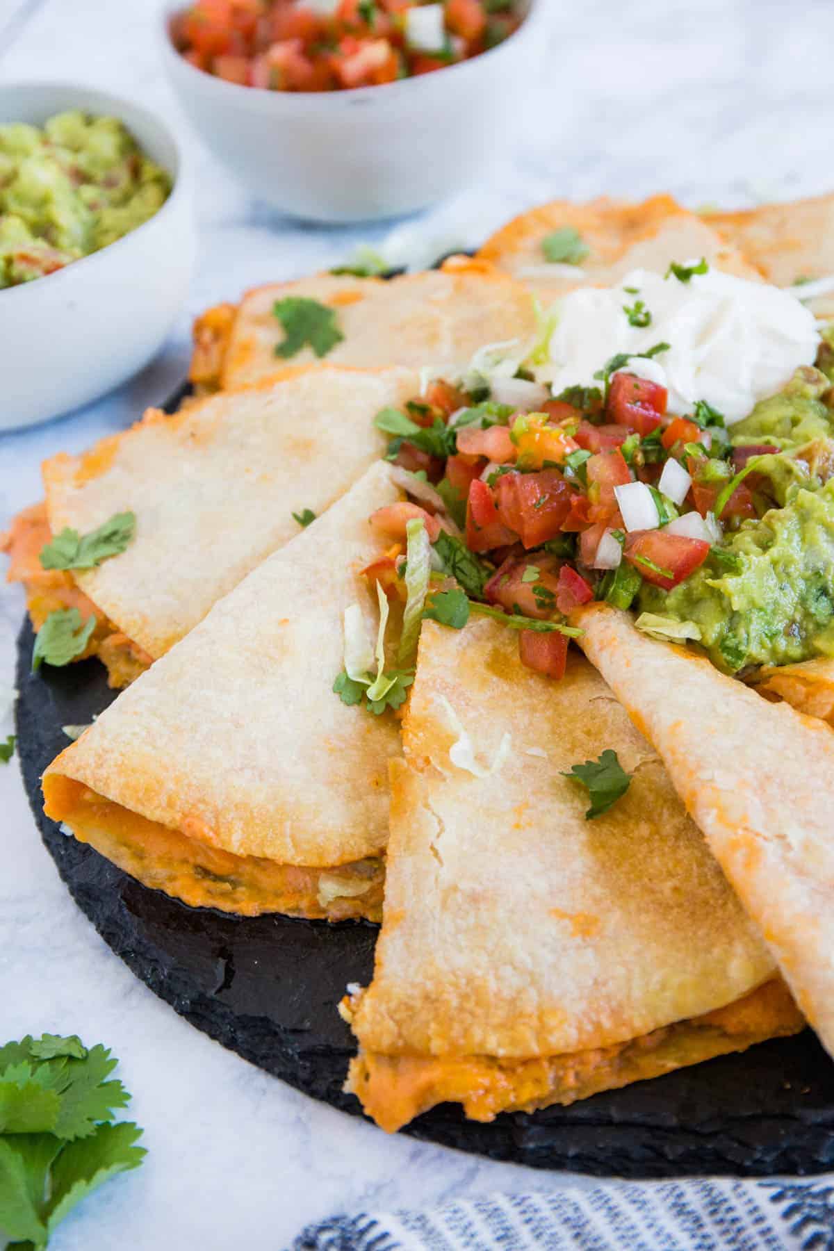 Cheesy quesadillas cut into wedges and served on a black plate, topped with fresh pico de gallo, guacamole, sour cream, and garnished with chopped cilantro. Bowls of guacamole and salsa are in the background.