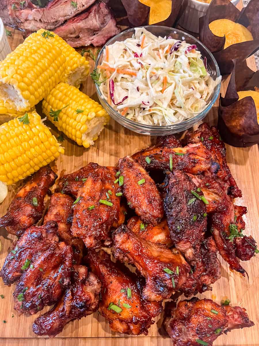 Barbecue chicken wings served on a wooden board with corn on the cob, creamy coleslaw in a glass bowl, and golden cornbread muffins. A colorful and hearty spread perfect for a game day or backyard party.
