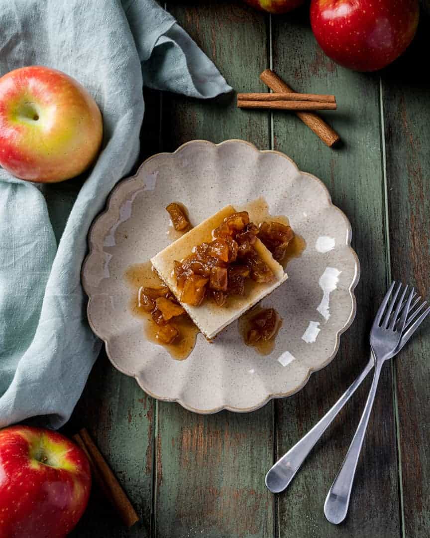 Apple cider cheesecake bars topped with caramelized apples, served on a scalloped plate with fresh apples, cinnamon sticks, and forks on a rustic green wood table.