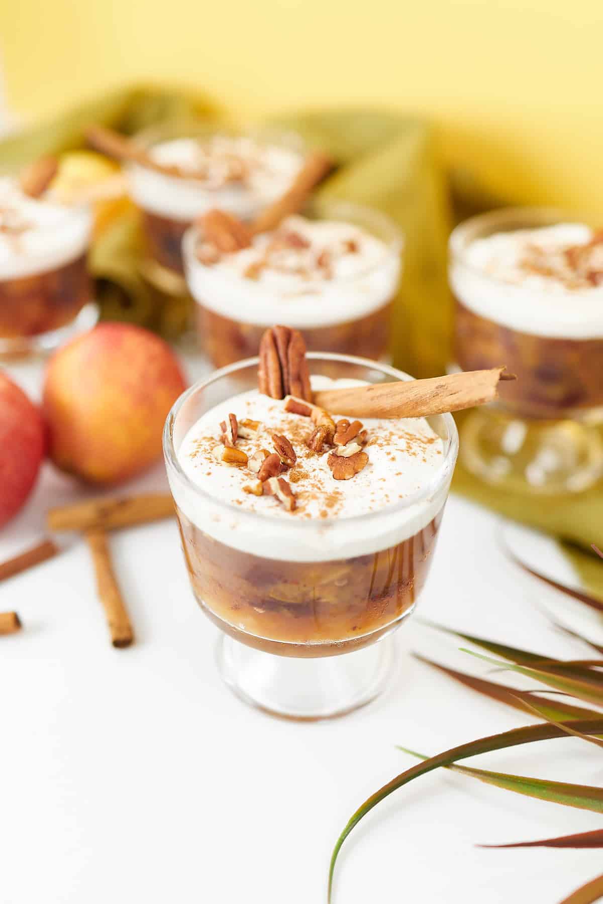 Individual servings of apple jello salad in glass dessert cups, topped with whipped cream, cinnamon, and chopped pecans, garnished with cinnamon sticks and surrounded by apples.