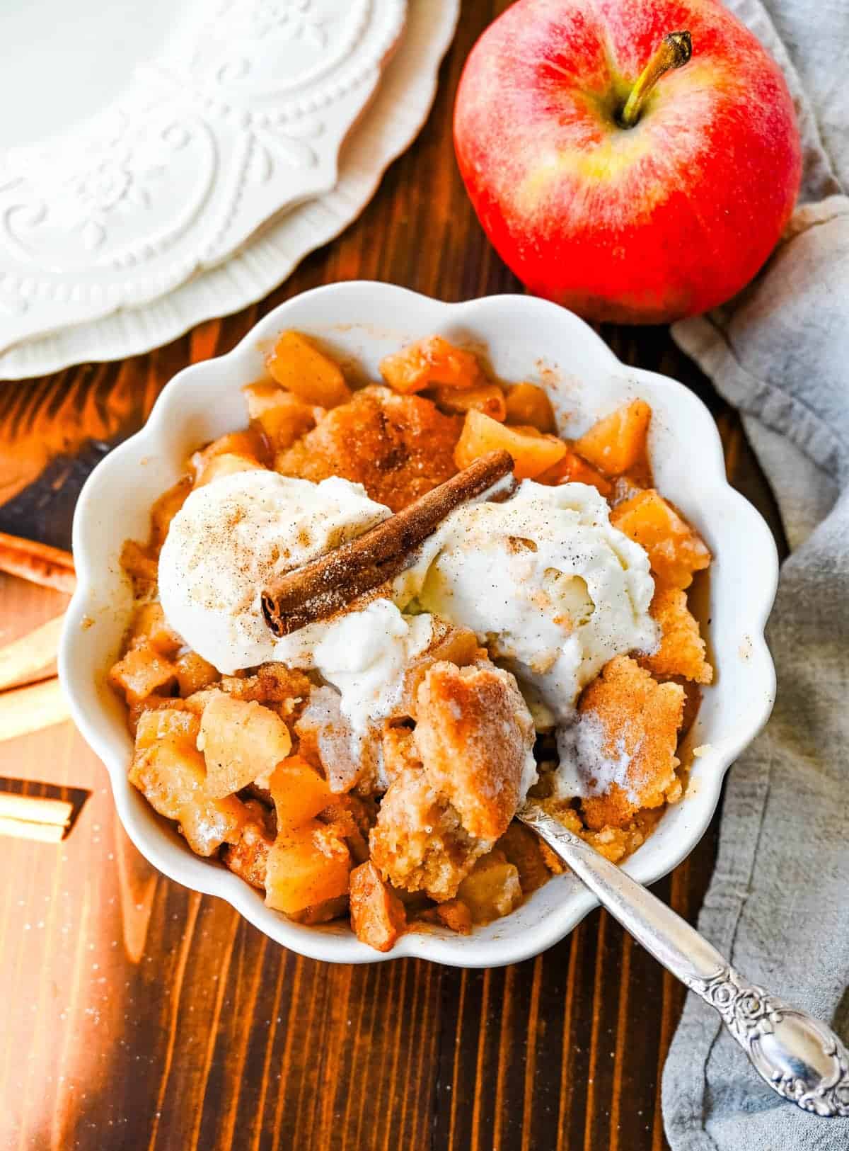 Warm apple cobbler served in a white scalloped bowl, topped with vanilla ice cream and a cinnamon stick, with chunks of cinnamon-spiced apples and a golden, crisp topping.