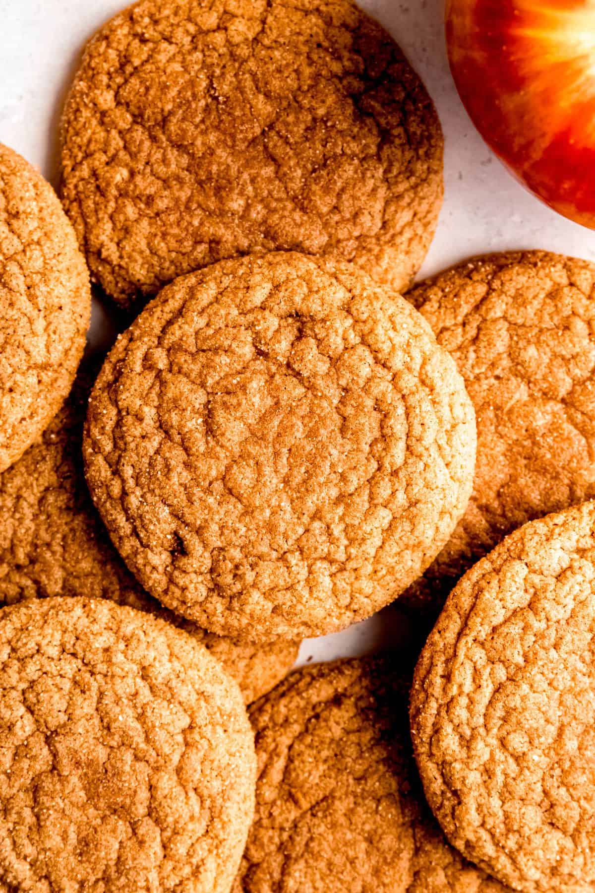 Close-up of golden brown cider cookies with a crackled cinnamon-sugar surface, arranged next to a red apple.
