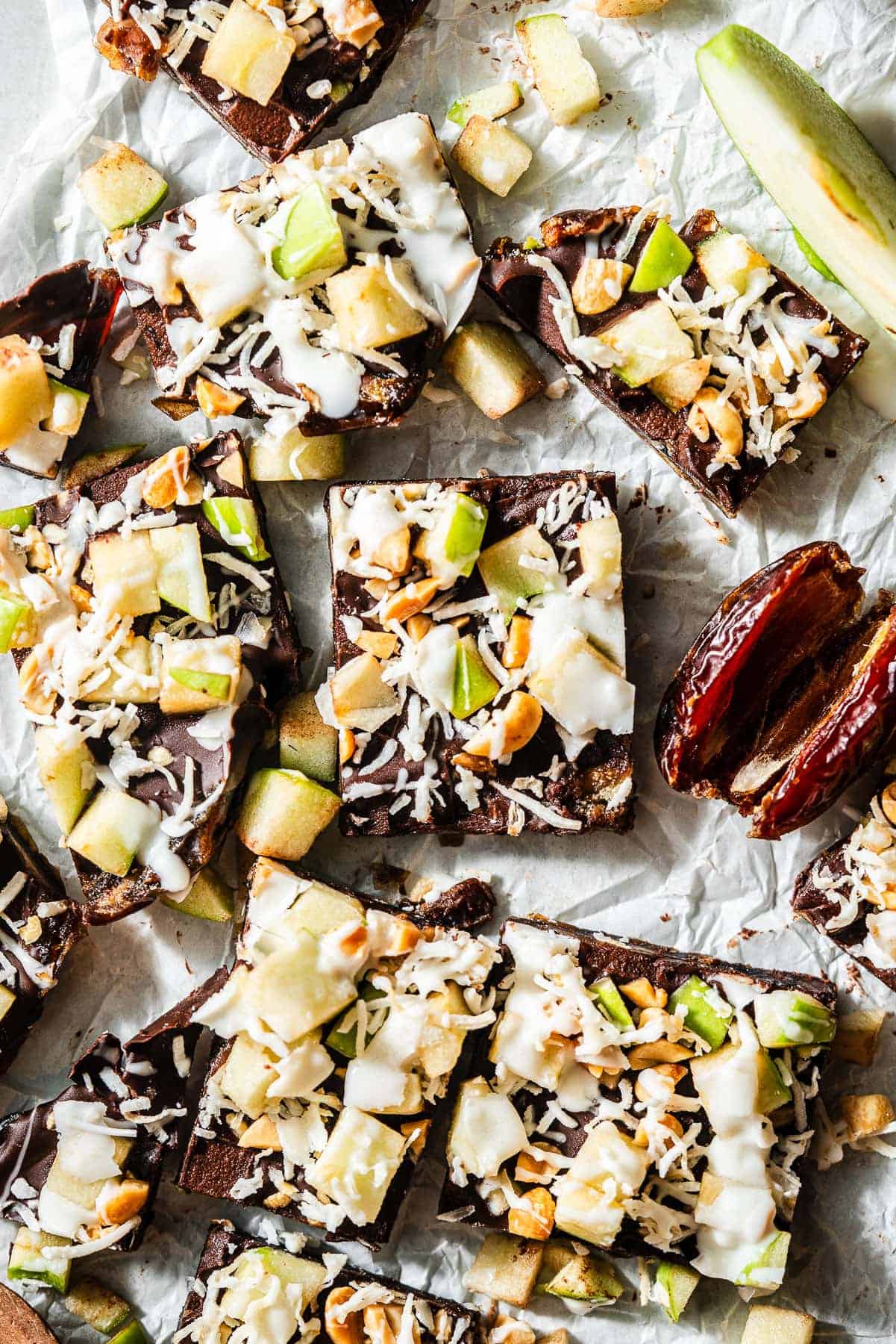 Squares of caramel apple date bark topped with chopped green apples, shredded coconut, cashews, and white drizzle on parchment paper with sliced dates nearby.
