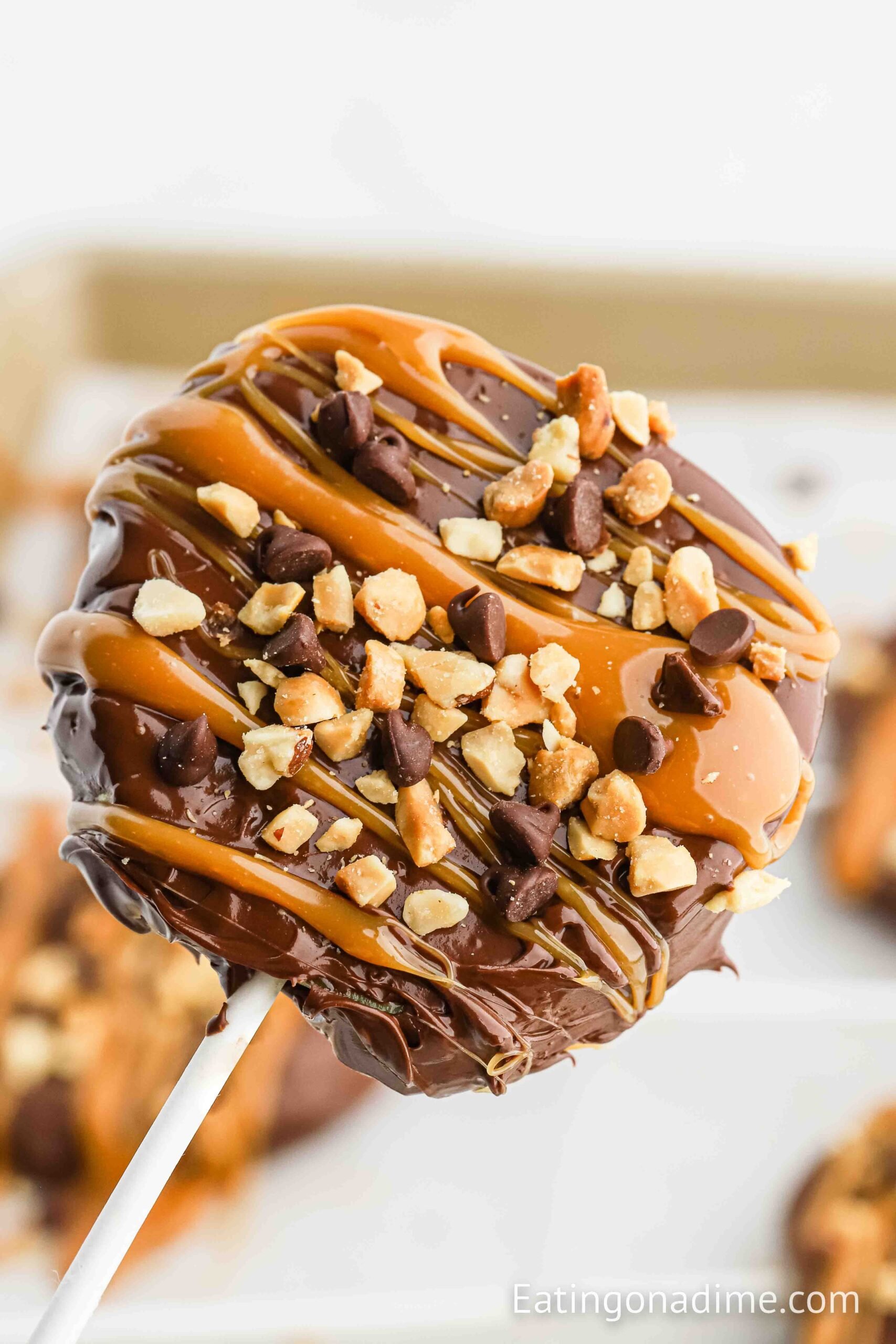 Close-up of a caramel apple slice on a stick, coated in chocolate and caramel, then topped with chopped nuts and mini chocolate chips.