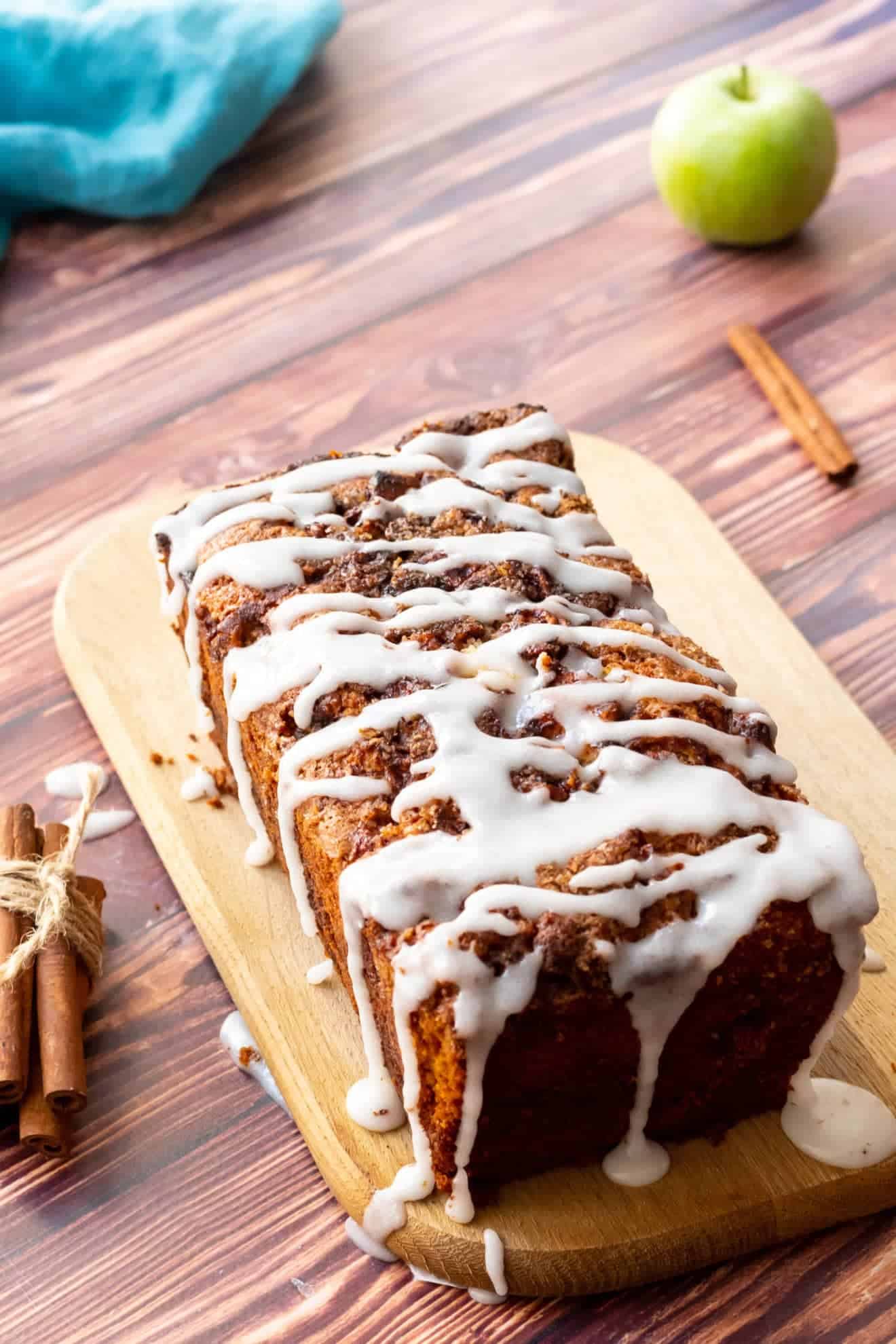 Loaf of apple fritter bread drizzled with vanilla glaze on a wooden board, with a green apple and cinnamon sticks in the background.