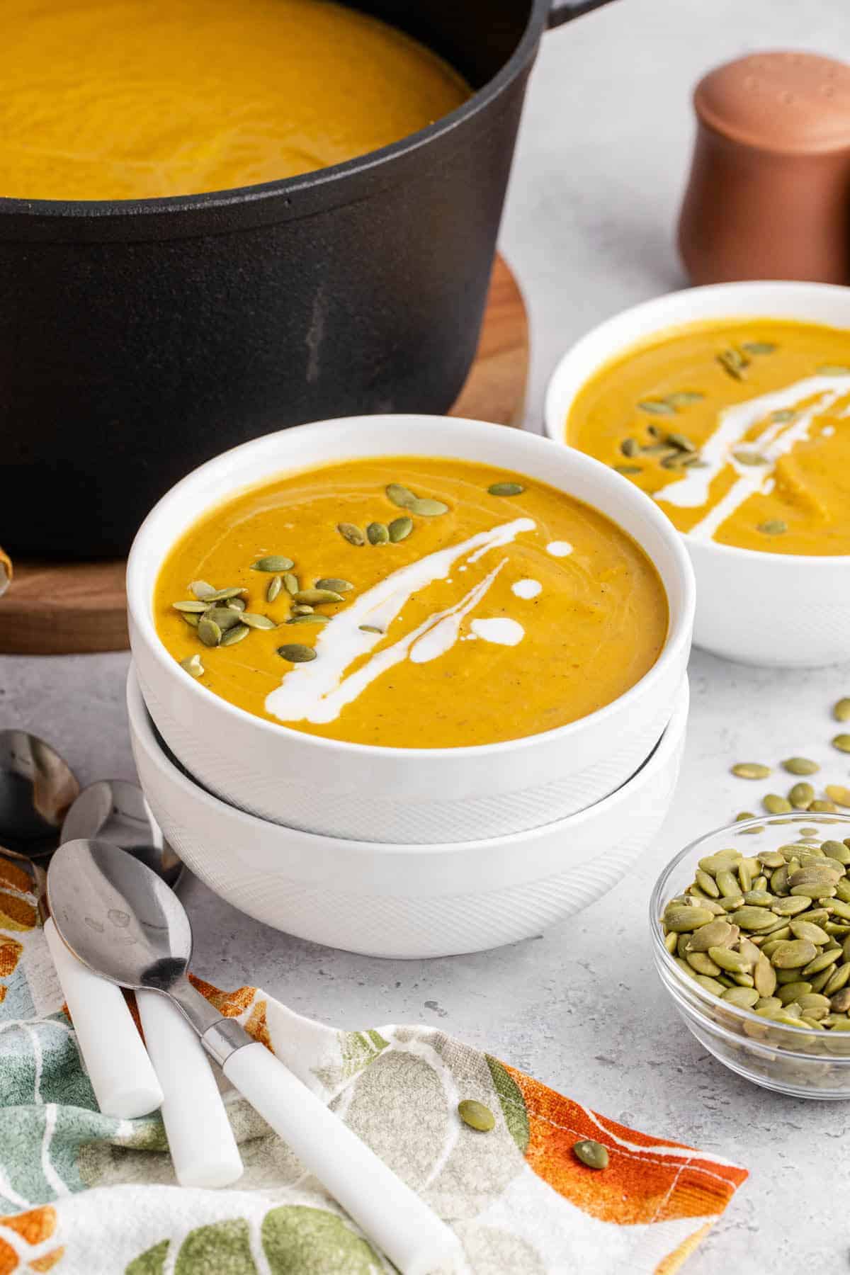 Creamy pumpkin carrot soup served in white bowls, topped with pepitas and a swirl of cream, with a pot of soup in the background.