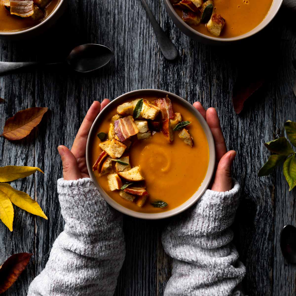 Bowl of pumpkin and sweet potato soup topped with crispy pancetta, sage, and croutons, held in hands over a rustic wooden table.