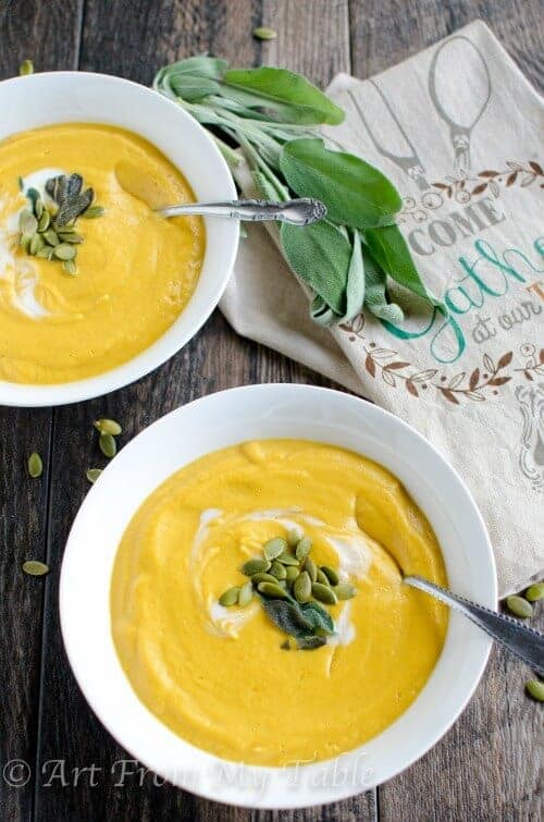Smoky pumpkin soup in white bowls topped with pepitas, sage, and cream swirl on a rustic wooden table.
