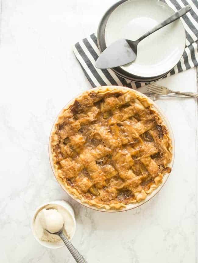 Golden brown vegan caramel apple pie with a lattice crust, served on a marble background next to a bowl of vanilla ice cream and pie-serving utensils.