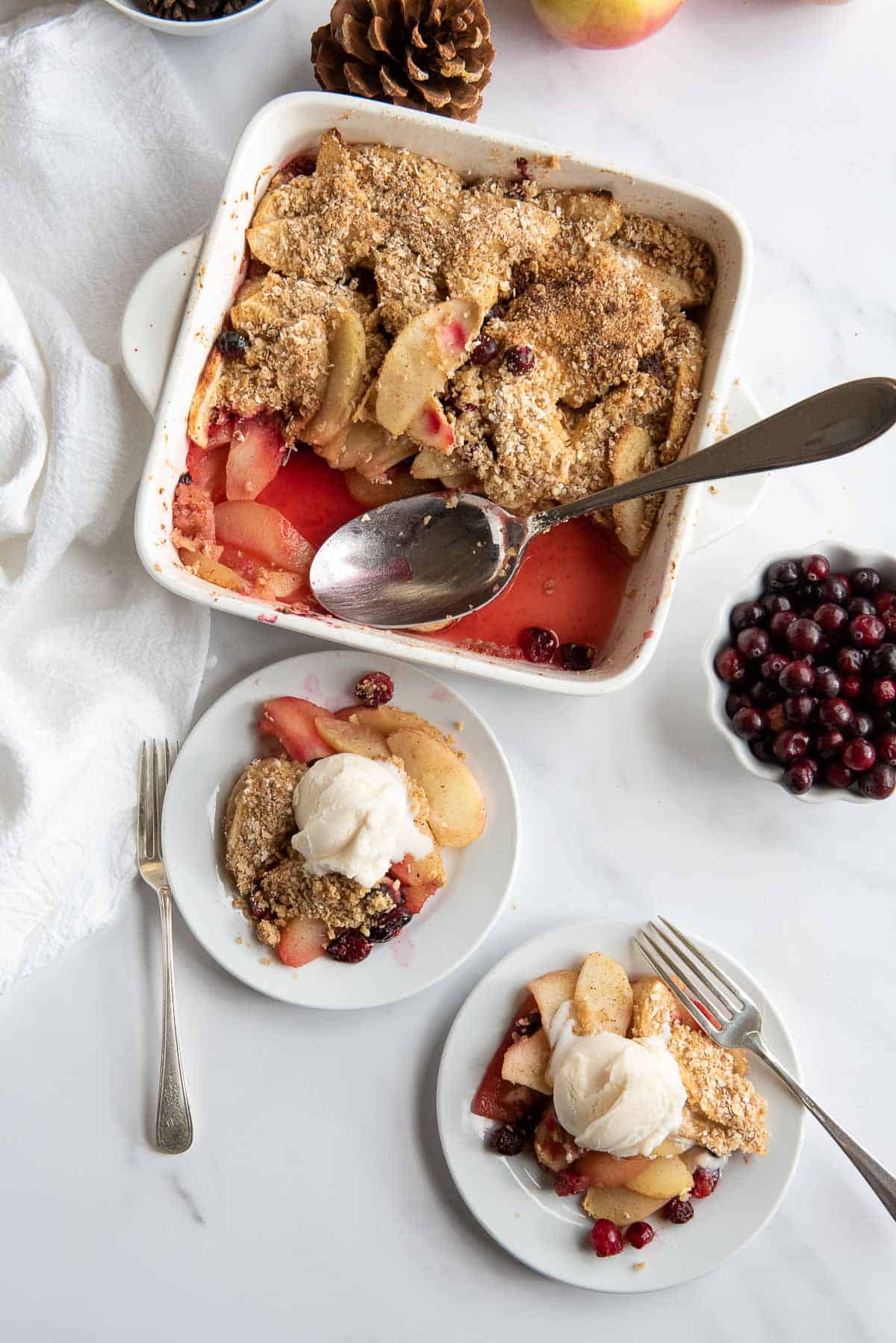 Baking dish filled with cranberry apple crisp, with two small plates of the dessert topped with vanilla ice cream on a white background, surrounded by fresh cranberries and pinecones.