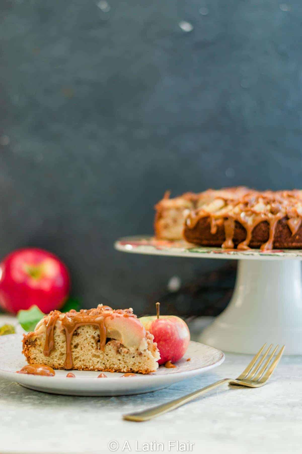 Slice of spiced apple streusel coffee cake drizzled with dulce de leche, served on a plate with a small apple, with the full cake on a stand in the background.
