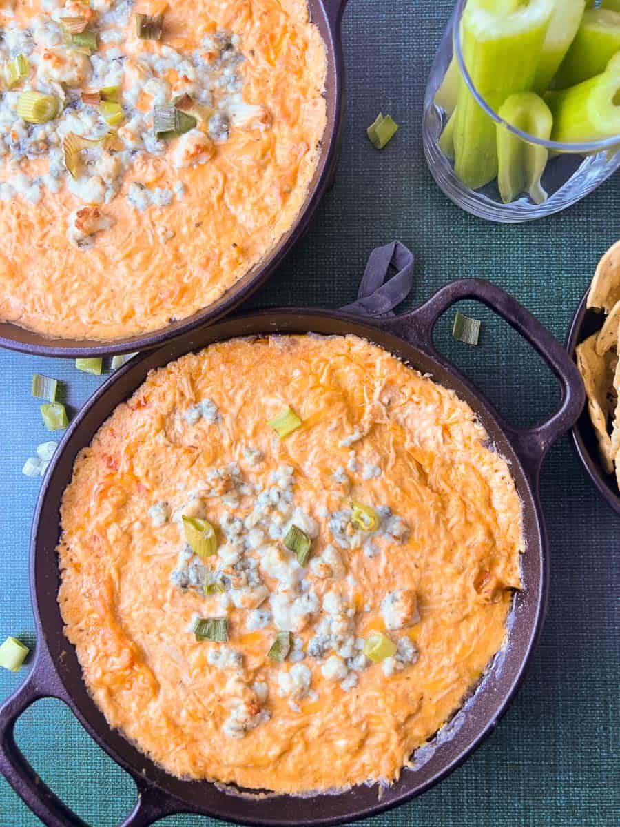 Buffalo chicken dip in a cast-iron skillet, hot from the oven, topped with melted cheese and green onions, served with celery sticks and tortilla chips perfect for game day or football snacks.