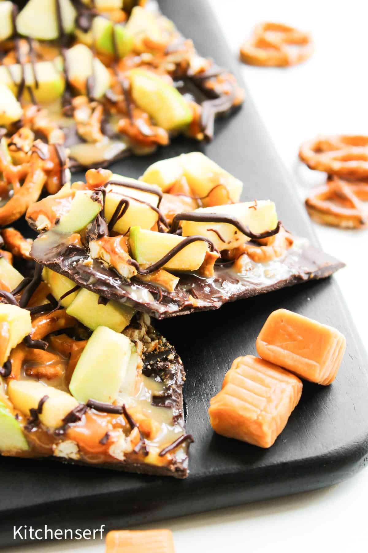 Close-up of caramel apple pretzel bark with chopped green apples, pretzel pieces, caramel drizzle, and dark chocolate on a black serving board with caramel candies and pretzels nearby.