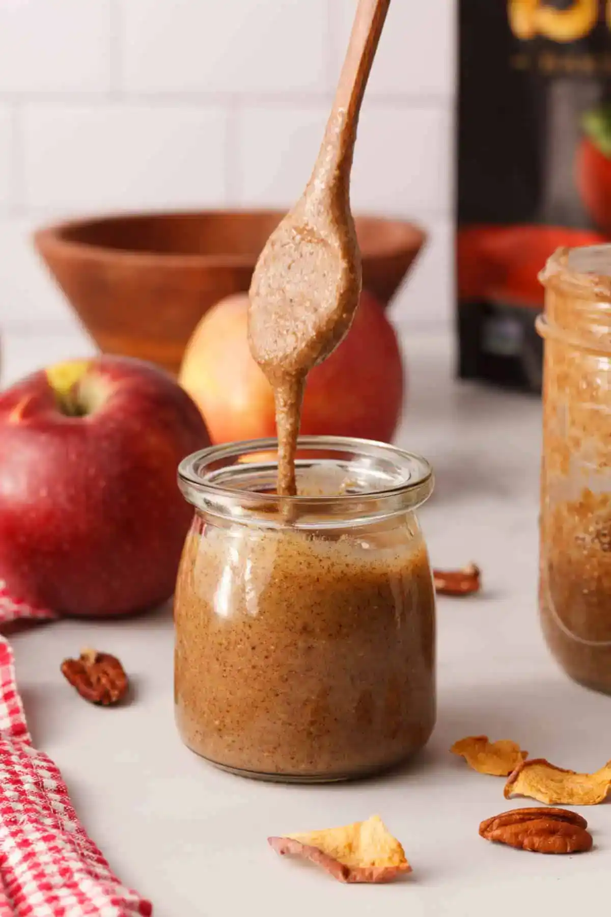 Jar of homemade apple pecan butter with a spoon drizzling the thick, creamy spread, surrounded by fresh apples, dried apple slices, and pecans on a white surface.