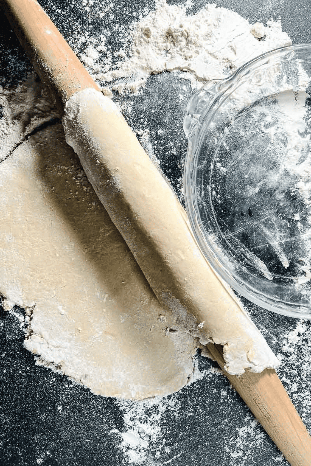 Rolled-out flaky pie crust dough on a floured surface with a wooden rolling pin and a clear glass pie dish nearby, showing the final step in preparing an easy homemade pie crust for baking a buttery, perfect pie crust.