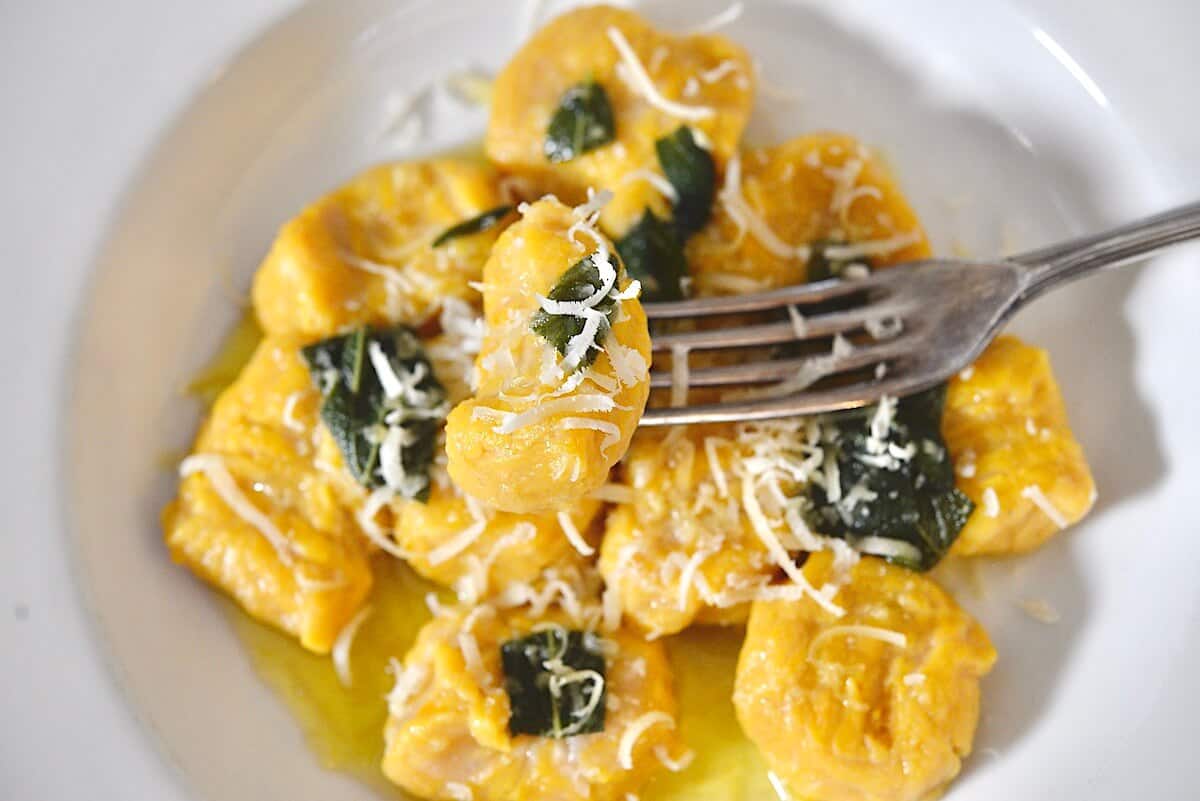 Homemade pumpkin gnocchi topped with crispy sage leaves, parmesan cheese, and browned butter on a white plate.