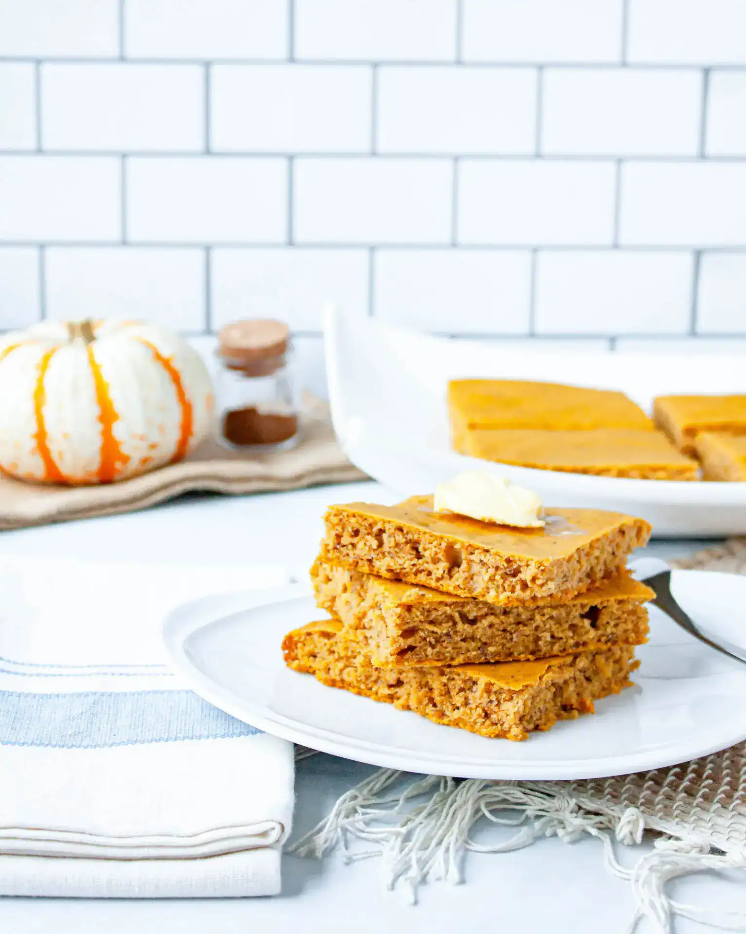 Pumpkin spice sheet pan pancakes stacked on a plate with butter on top, served alongside fall decor and a tray of baked pancakes.