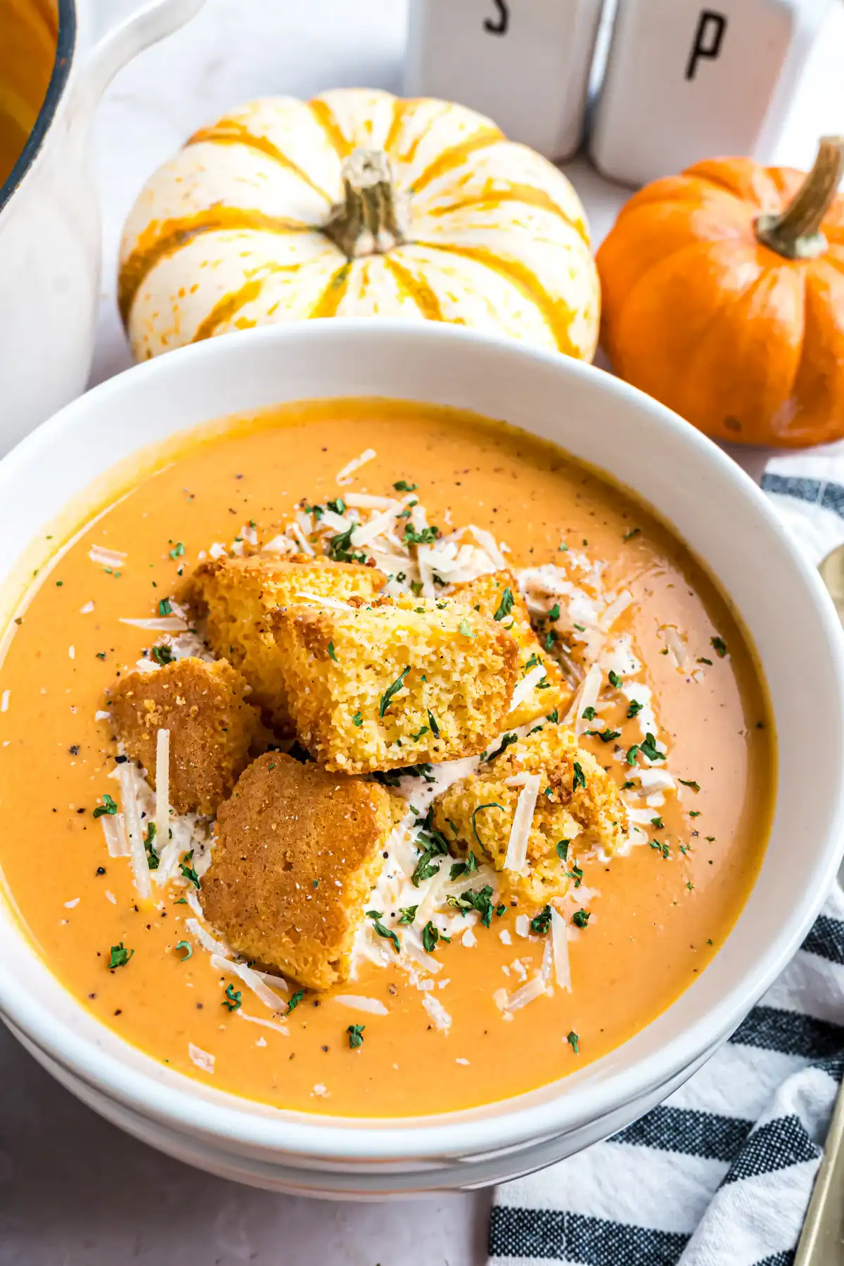 Creamy pumpkin tomato soup topped with croutons, parmesan cheese, and herbs in a white bowl with small pumpkins in the background.