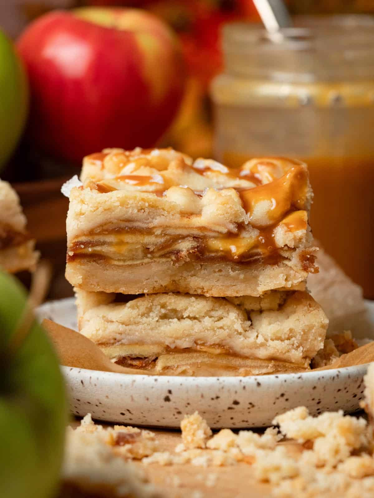 Stacked caramel apple pie bars with a buttery shortbread crust and caramel drizzle, served on a speckled plate with fresh apples and a jar of caramel in the background.