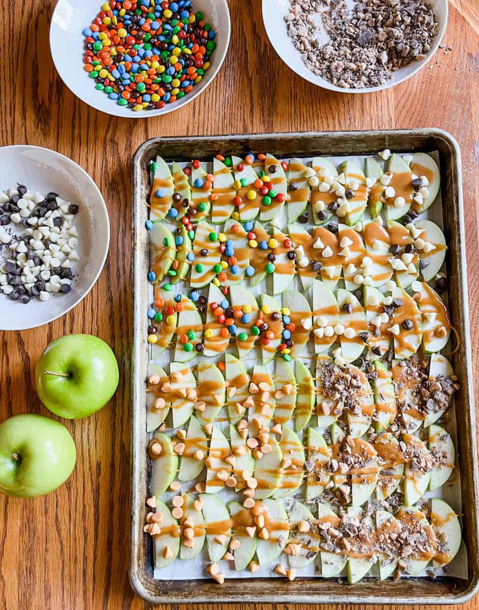Sheet pan loaded with sliced green apples drizzled with caramel sauce and topped with various candy toppings including M&Ms, white and dark chocolate chips, toffee bits, and peanut butter chips, surrounded by bowls of toppings on a wooden table.