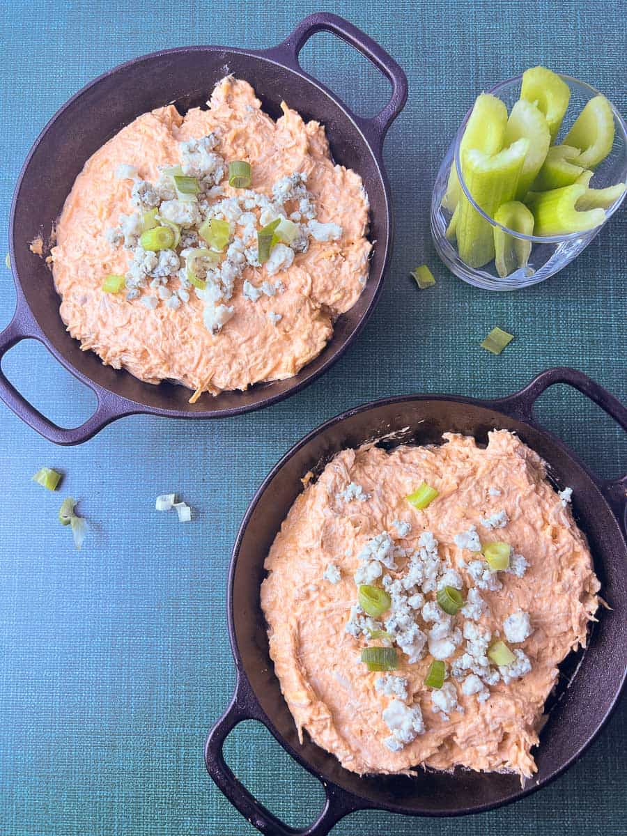 Two cast iron skillets filled with creamy buffalo chicken dip topped with crumbled blue cheese and sliced green onions, served with a glass of fresh celery sticks for the perfect easy buffalo chicken dip and game day food.