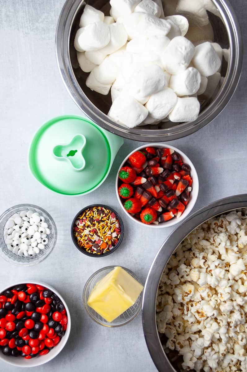 Ingredients for making Halloween marshmallow popcorn balls, including a large bowl of marshmallows, popped popcorn, a stick of butter, Halloween sprinkles, candy eyes, red and black M&M’s, and festive candy corn pumpkins. A green popcorn ball mold is also shown, perfect for shaping this no-bake Halloween treat.
