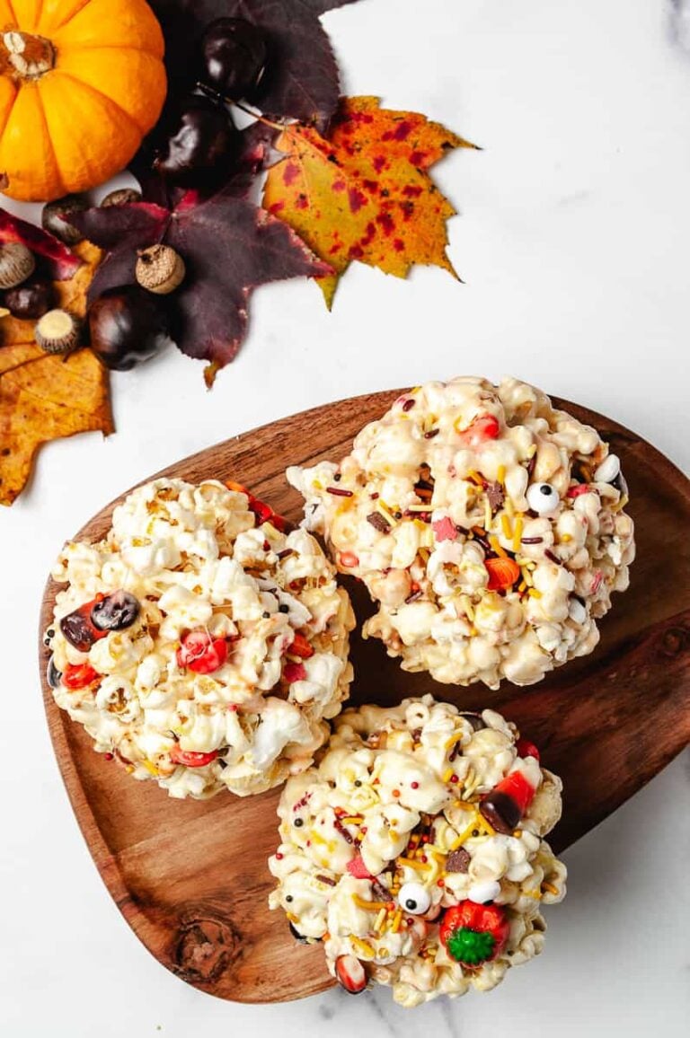 Three gooey Halloween marshmallow popcorn balls decorated with colorful Halloween sprinkles, candy eyes, and M&M's sit on a wooden tray. Surrounded by fall leaves, acorns, and a small pumpkin, this no-bake Halloween dessert makes the perfect festive treat for spooky season or kids’ Halloween parties.
