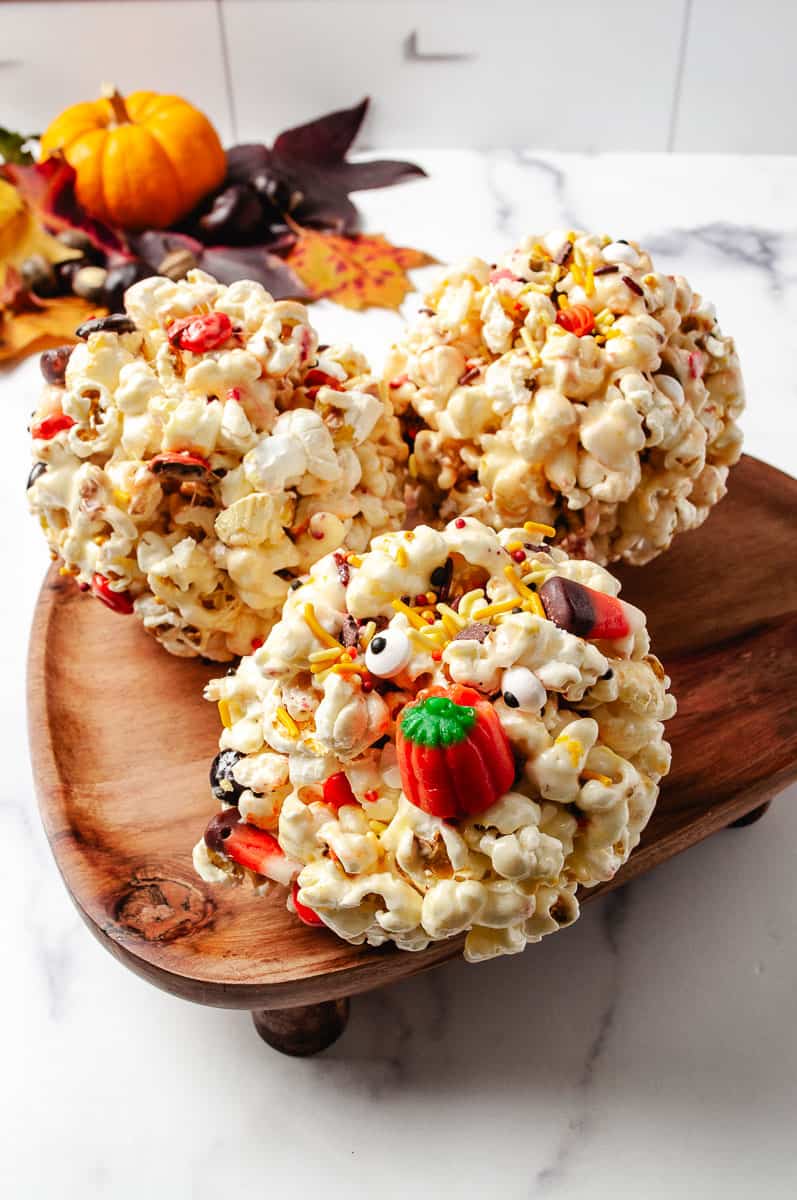 Three gooey Halloween marshmallow popcorn balls decorated with candy pumpkins, candy eyes, Halloween sprinkles, and colorful chocolate candies, displayed on a wooden tray with fall leaves and a mini pumpkin in the background. A fun, no-bake Halloween treat perfect for spooky season and Halloween parties for kids.