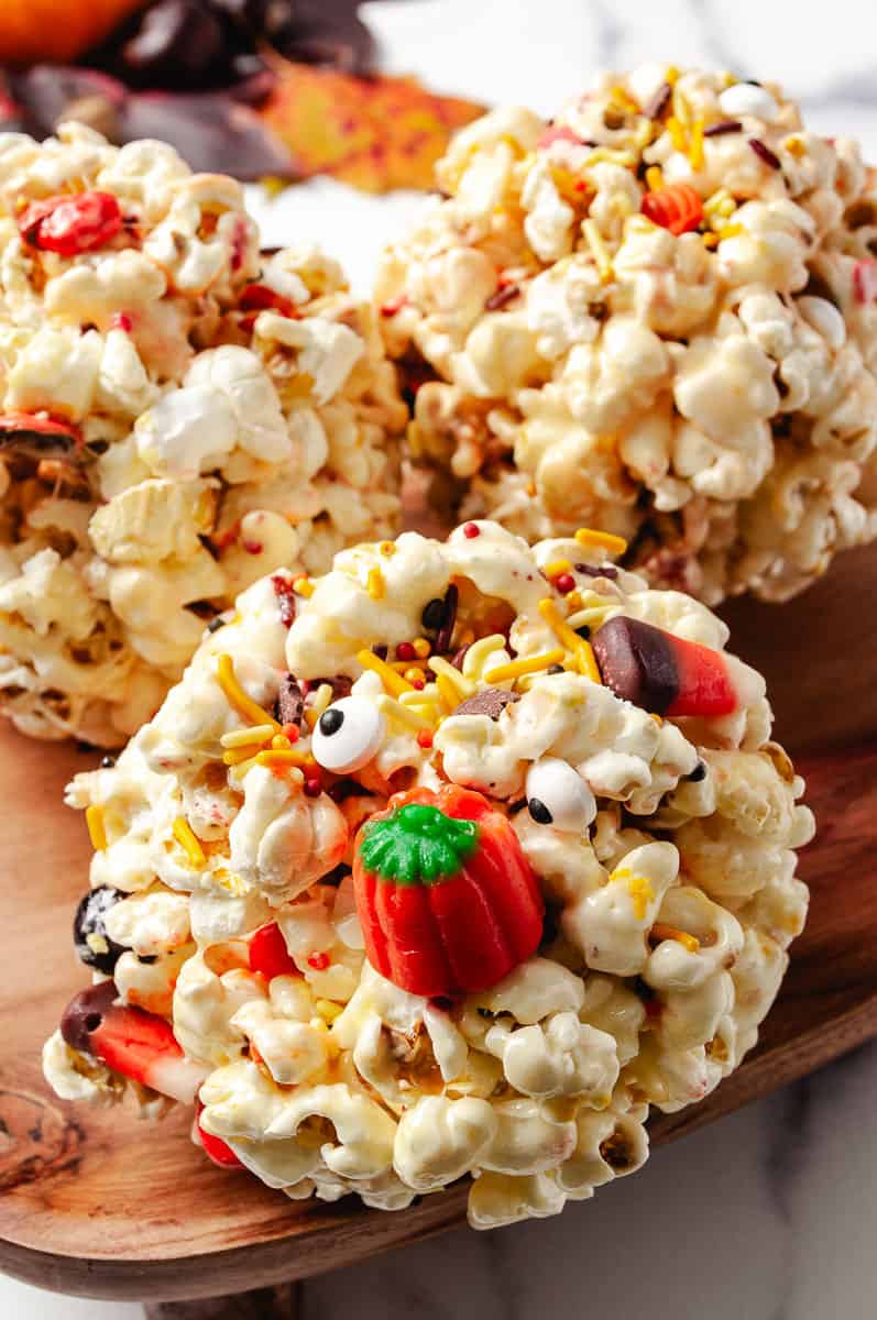 Close-up of a gooey Halloween marshmallow popcorn ball topped with candy eyes, Halloween sprinkles, and a gummy pumpkin, with two more popcorn balls in the background. This no-bake Halloween treat is perfect for spooky season parties and kid-friendly fall snacks.