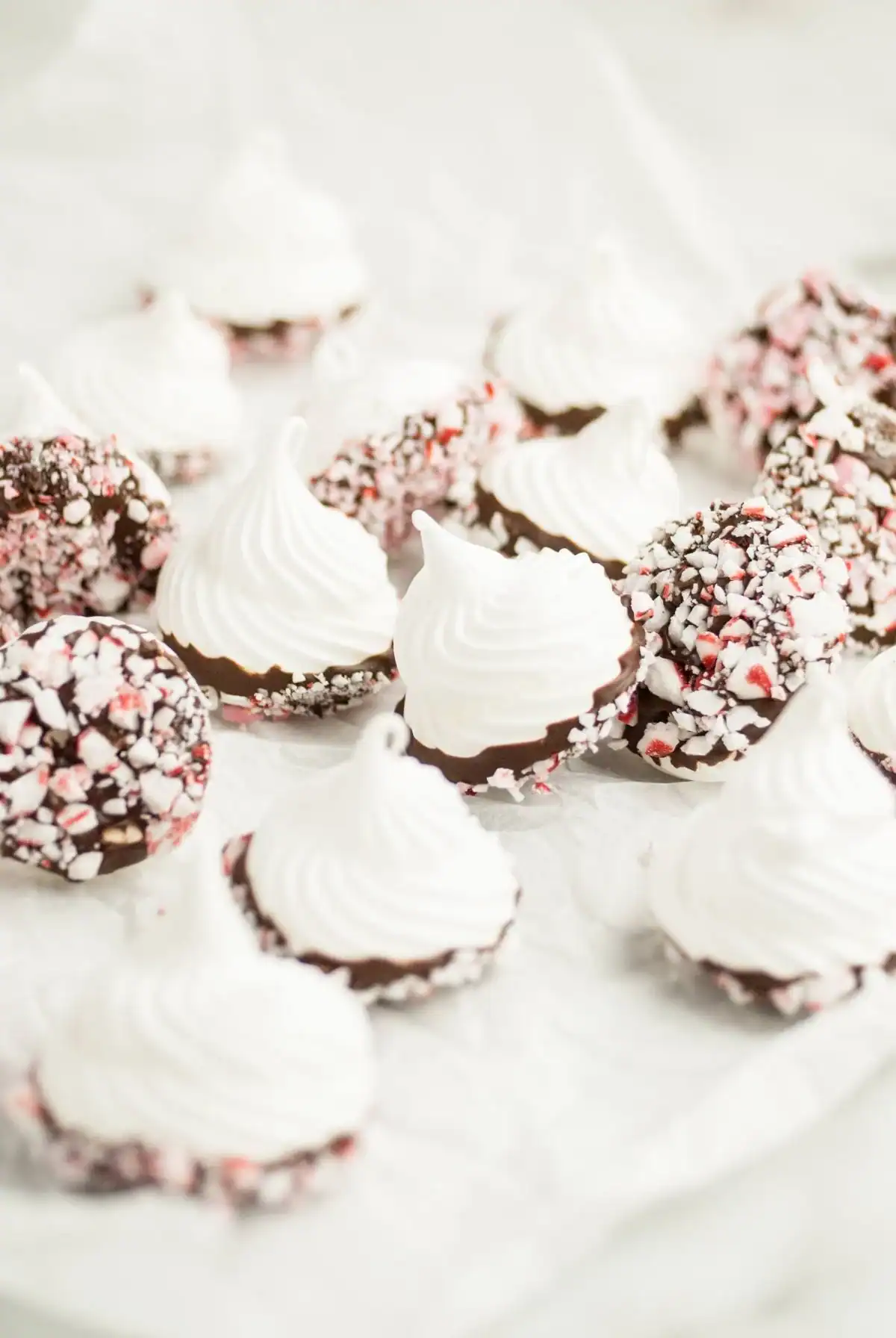Chocolate dipped peppermint meringues with crisp white tops and bases coated in chocolate and crushed candy canes, arranged on parchment paper for the holidays.