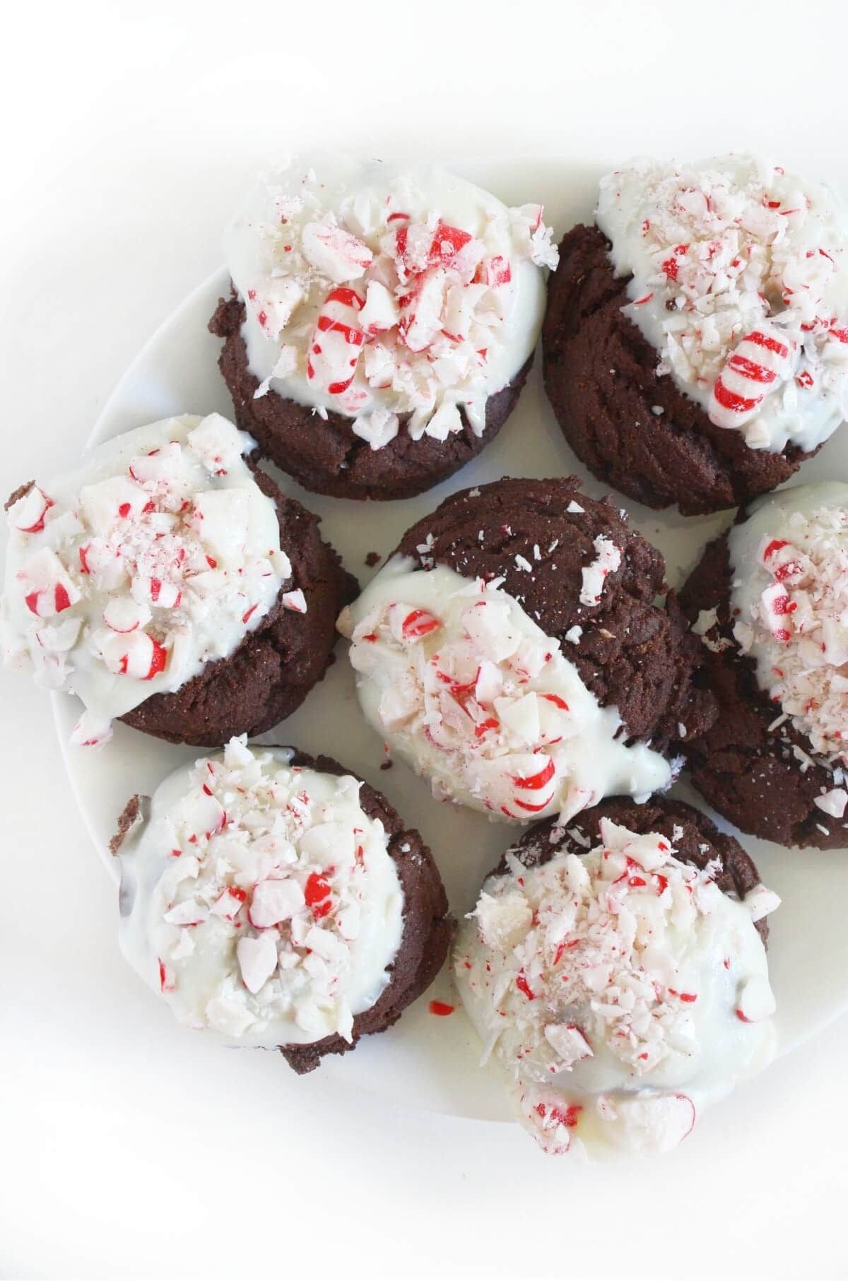 Gluten-free, vegan peppermint bark cookies with fudgy chocolate bases, topped with white chocolate and crushed candy canes, displayed on a white plate.