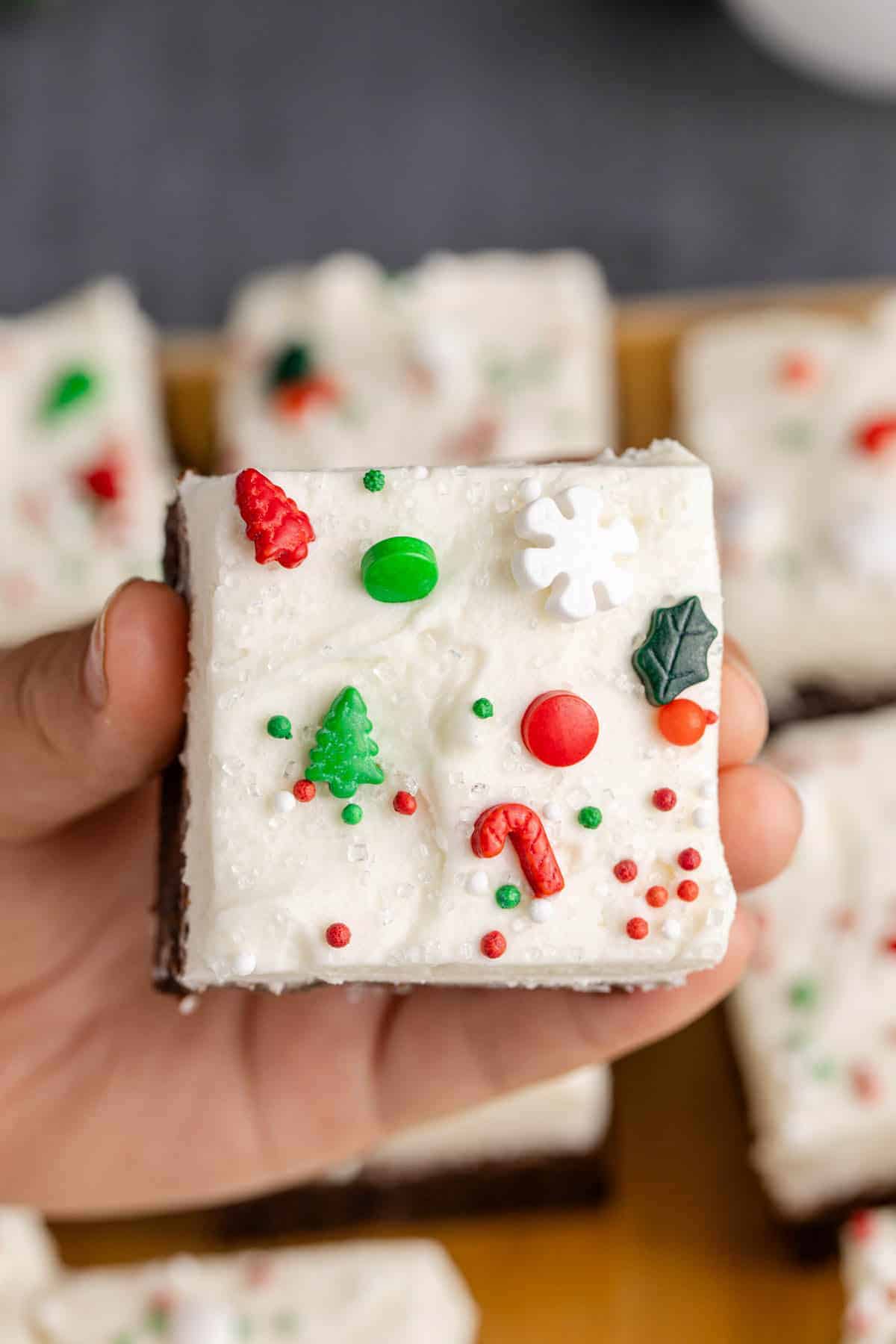 Frosted peppermint brownie topped with creamy white icing and colorful Christmas sprinkles including trees, candy canes, and snowflakes, held in hand.